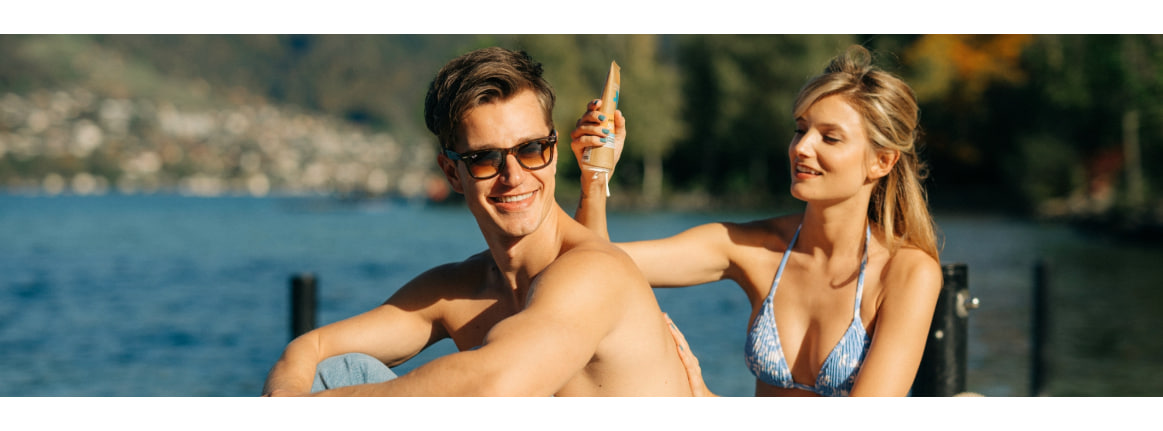 mann und frau am see. frau cremt mann den rücken ein.