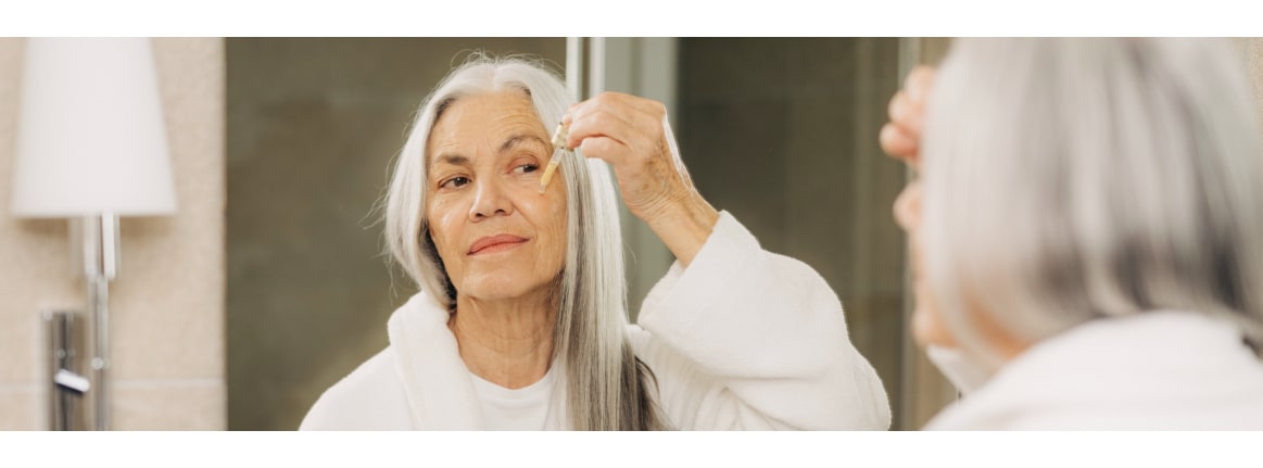 ältere frau mit langen grauen haaren tropft mit einer pipette serum auf ihr gesicht